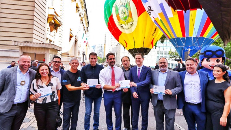 Foto posada. Leite ao centro e dez pessoas ao seu lado. Alguns deles seguram uma espécie de certificado nas mãos. Ao fundo o Palácio Piratini e dois balões, sendo um deles nas cores da bandeira do RS e com o brasão do Estado bem grande estampado.