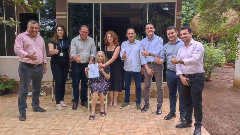 Nove pessoas (três mulheres e seis homens) posam para a câmera, sorrindo. Ao centro, sentada, está a aposentada Vilma Borelli, segurando um documento. As demais pessoas estão em pé, alinhadas atrás da senhora.