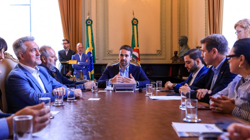 Foto em ambiente fechado. Ao redor de uma mesa retangular, estão sentadas diversas autoridades. Na ponta da mesa, está o governador Eduardo Leite falando e gesticulando. Ao fundo, aparecem as bandeiras do Brasil e do Rio Grande do Sul. Sobre a mesa, em frente às pessoas participantes da reunião, há copos de água e papéis.