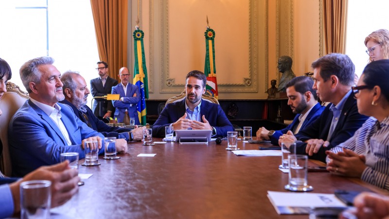 Foto em ambiente fechado. Ao redor de uma mesa retangular, estão sentadas diversas autoridades. Na ponta da mesa, está o governador Eduardo Leite falando e gesticulando. Ao fundo, aparecem as bandeiras do Brasil e do Rio Grande do Sul. Sobre a mesa, em frente às pessoas participantes da reunião, há copos de água e papéis. 