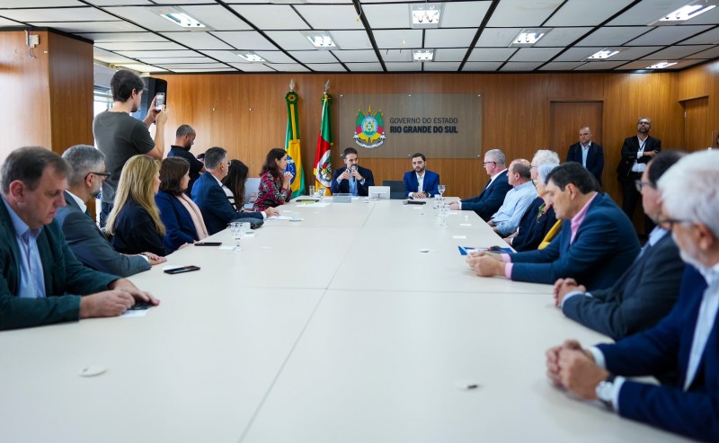 Em uma grande mesa de reunião, Leite e Gabriel estão na cabeceira e são vistos mais ao fundo. Leite fala ao microfone. Os participantes da reunião, sentados à direita e à esquerda, direcionam a atenção para o governador enquanto ele fala.