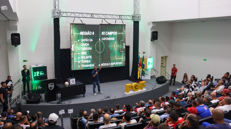Imagem mostra um auditório lotado. À frente, em cima do palco, há um homem apresentando informações sobre a Copa RS. Há um telão iluminado, com as seguintes palavras escritas em branco: Região 8: Capão da Canoa, Cidreira, Imbé, Rolante, Torres e Tramandaí. 01 Campos: Caponense e Módulo Esportivo. A imagem de fundo do telão é um campo de futebol. As pessoas na plateia estão olhando para o palco. 