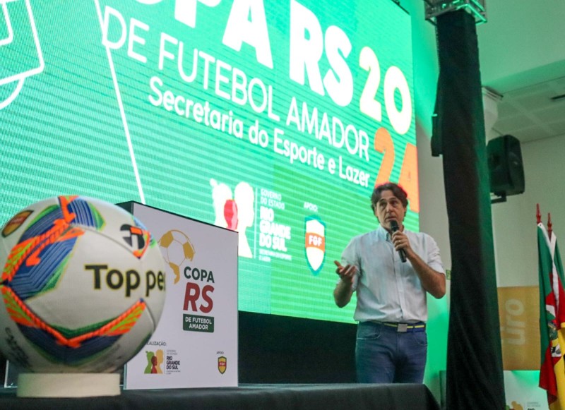 Imagem do titular da SEL, falando ao microfone. Ele está de pé, em frente a um telão iluminado, em tons verdes. No telão, há a marca do governo do Rio Grande do Sul e da FGF, além das palavras em branco "Copa RS 2024 de Futebol Amador - Secretaria do Esporte e Lazer".