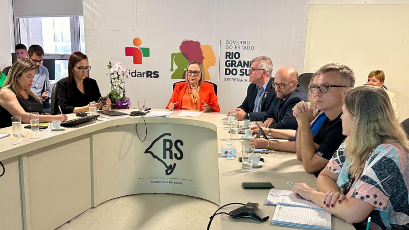 Em uma mesa de reunião em forma de U estão sentados a secretária da Saúde e alguns representantes da saúde discutindo o Plano Estadual de Contrarreferência.