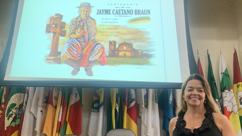 Cíntia aparece sorrindo, de pé, olhando para a câmera. Ao fundo, num slide, está a identidade visual, que consiste num desenho de Jayme sentado, segurando um chimarrão e com roupas tradicionais; à esquerda dele está uma cruz, e ao fundo uma construção das Missões.