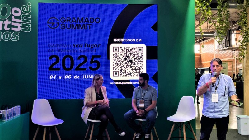 Um homem mais à direita em pé falando ao microfone e dois outros participantes sentados mais ao centro. Atrás deles um painel no qual está escrito Gramado Summit e um QR code.