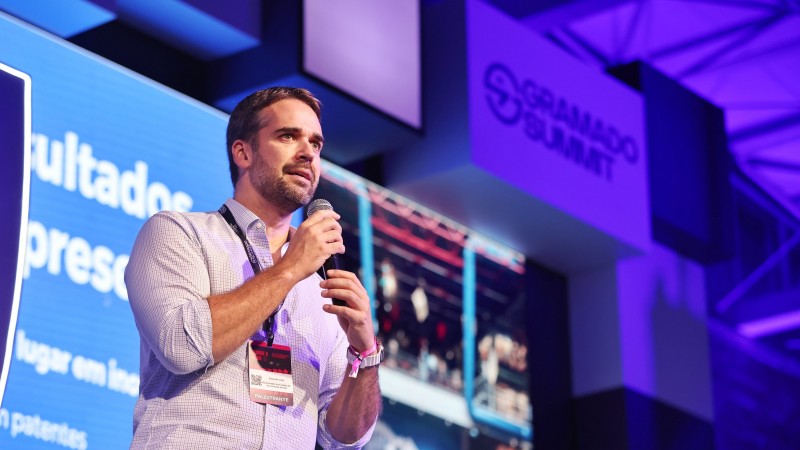Foto do governador Eduardo Leite. Ele está falando ao microfone e usando o crachá do Gramado Summit. Ao fundo, há um painel iluminado e a marca do evento.