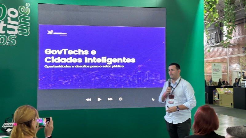 Foto da palestra de Ayub, que aparece de pé falando ao microfone. Ao fundo, há um grande painel verde com uma tela na qual está escrito "Govtechs e Cidades Inteligentes".