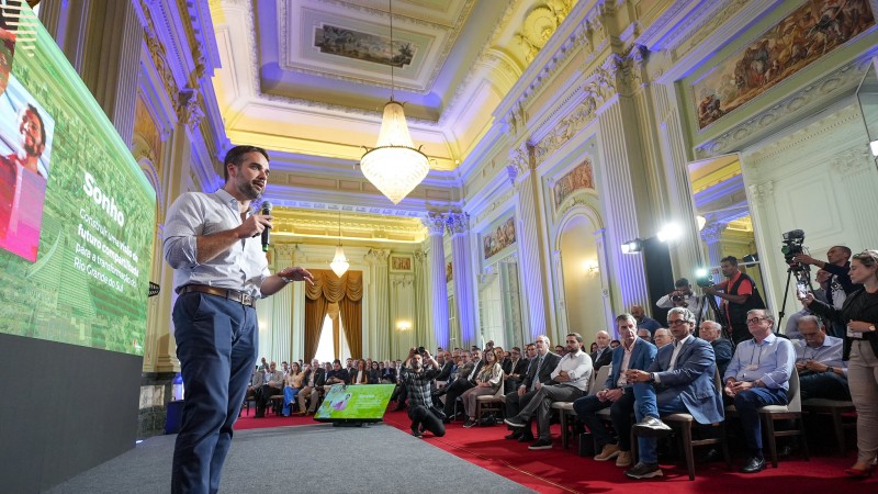 Imagem em ambiente fechado, no Palácio Piratini. Em destaque, posicionado à esquerda da foto, está o governador Eduardo Leite, falando ao microfone. Ao fundo, há um painel com a apresentação. Do lado direito da foto, estão diversas pessoas na plateia, escutando a fala do governador. 