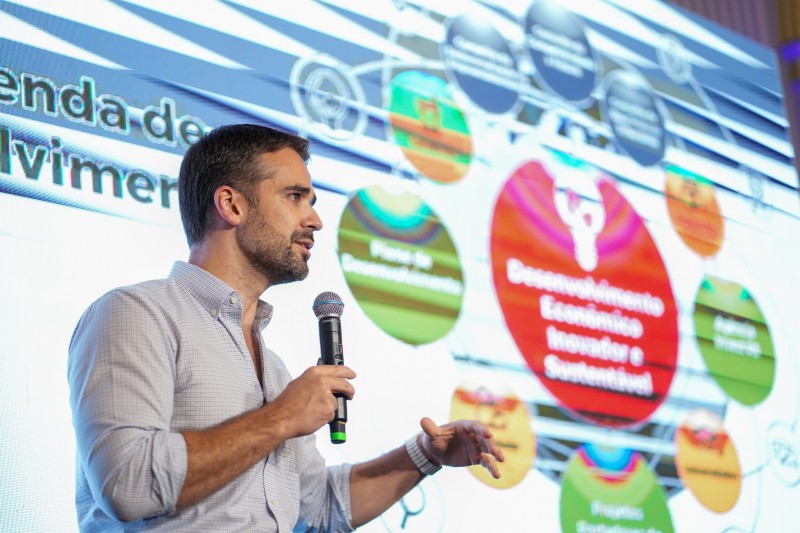 Foto do governador Eduardo Leite falando ao microfone e gesticulando. Ele está posicionado diante de um painel com a apresentação.
