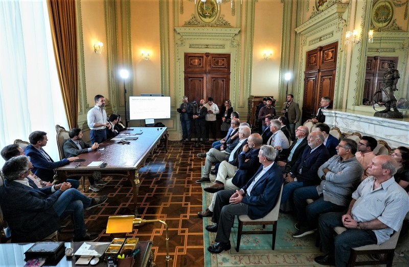 Reunião com entidades sobre sustentabilidade fiscal do Estado. Todos de perfil. Leite, à esquerda da imagem, está em pé atrás de uma mesa à qual estão sentados outros cinco homens. Ele fala com a plateia que está mais à direita da imagem. Ao fundo e mais ao centro podem ser vistas algumas pessoas que fazem a cobertura da reunião.