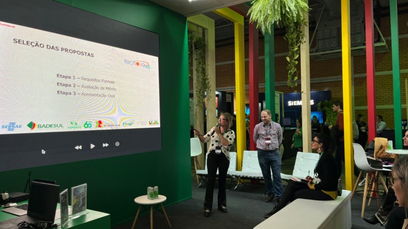 Foto em ambiente fechado de um homem e uma mulher de pé apresentando o painel. A mulher fala ao microfone enquanto olha para o slide ao fundo. Há bancadas brancas no meio e pilastras coloridas ao fundo.
