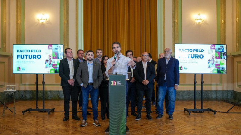 Foto em ambiente interno, no Palácio Piratini. Leite está à frente de um grupo de autoridades, falando ao microfone e gesticulando. Todos estão em pé, posicionados próximos um ao outro. Do lado esquerdo e direito da foto há painéis iluminados, onde se lê: "Pacto pelo futuro", a marca do governo do Estado e fotografias de pessoas.