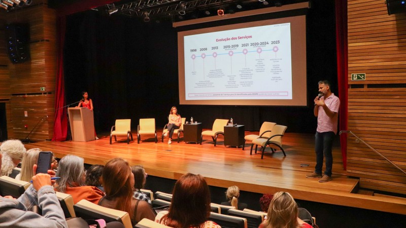 Foto em um auditório amadeirado. Um homem de pé fala ao microfone, no palco. Há cadeiras, e uma mulher se senta numa dela. Há também uma mulher junto a um púlpito. Ao fundo, há uma apresentação de slides. Num canto inferior da foto aparecem pessoas na plateia.