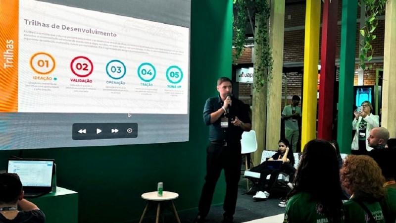 Rosa em pé ao microfone falando à plateia. Em uma tela estão projetados números que correspondem ao conteúdo oferecido às startups na plataforma. Podem ser vistas algumas pessoas da plateia de costas assistindo à apresentação e no canto inferior esquerdo o operador da apresentação projetada.