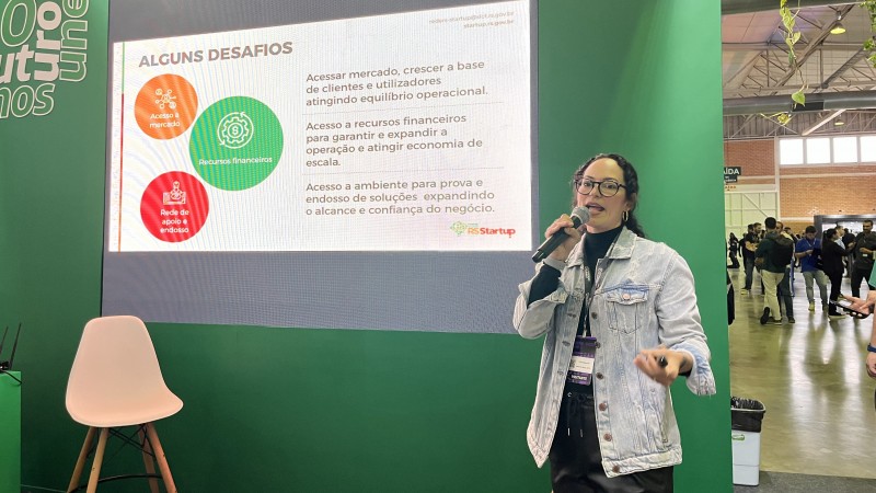 Foto em plano americano de Emily, de pé, falando ao microfone durante o evento. Ao fundo, há um grande painel verde com o slide de apresentação.