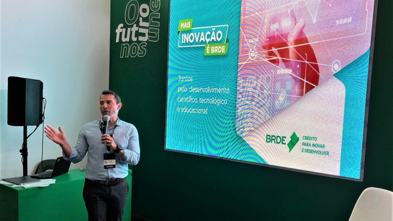 Leonardo Busatto está em pé e falando ao microfone mais à esquerda da imagem. Mais à direita, há um painel onde se vê uma projeção que diz: Mais inovação é BRDE.
