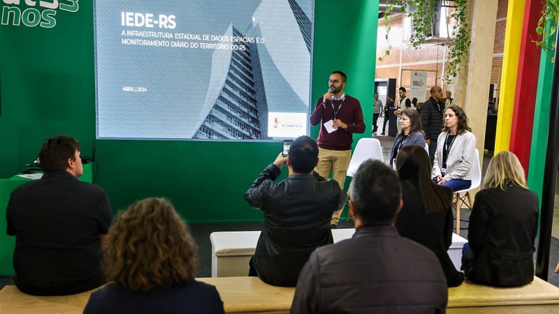 Alguma pessoas sentadas na plateia. Elas aparecem de costas na imagem assistindo à apresentação de um homem que está em pé e com o microfone diante da plateia. Na tela está projetado; IEDE-RS. As outras duas participantes do painel são vistas sentadas à direita do palestrante.