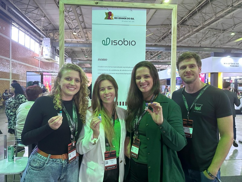 Foto posada de três mulheres e um homem no evento. Todos estão de pé e sorriem. Ao fundo, há um banner com o nome da startup.