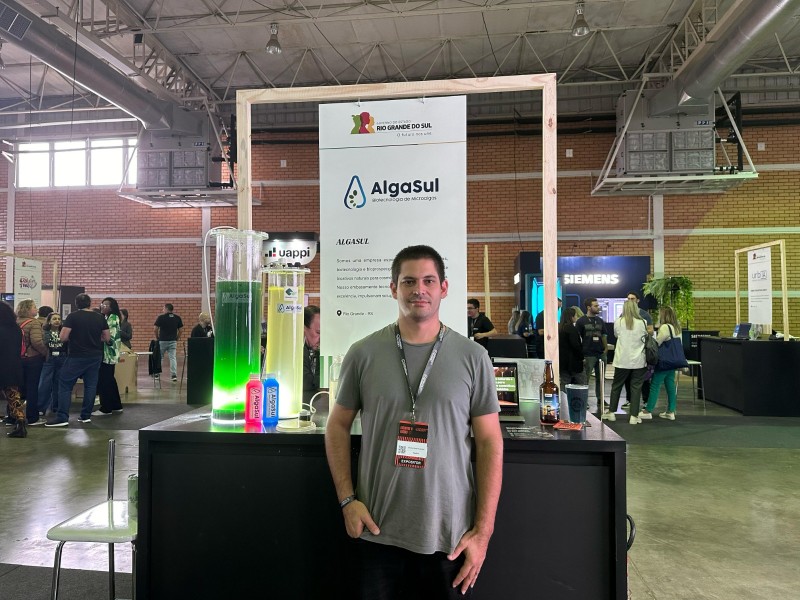 Foto posada de Bruno de pé, olhando para a câmera. Ao fundo, um banner com o nome AlgaSul. Atrás, há pessoas na feira.