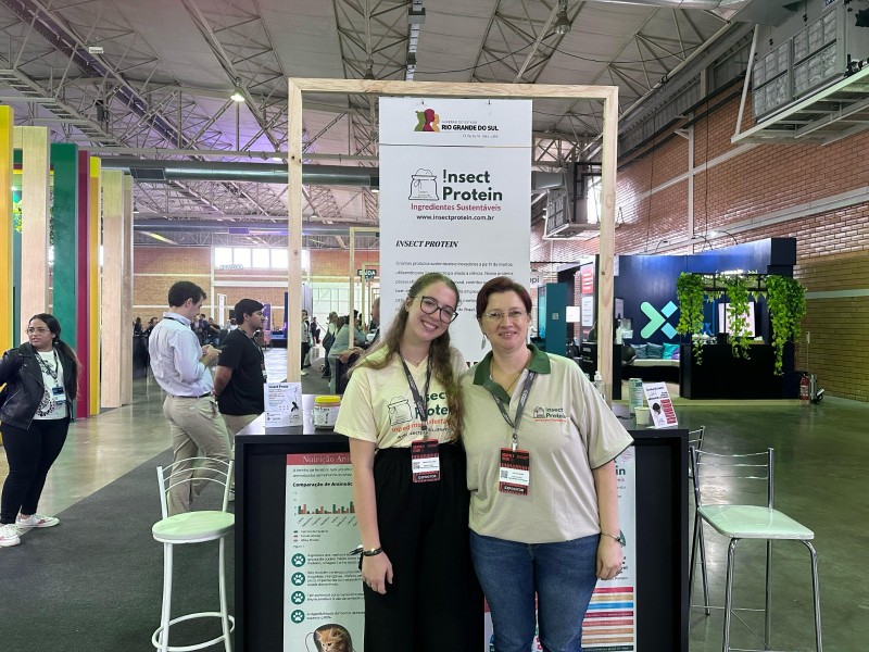 Foto posada de duas mulheres de pé, sorrindo. Elas usam camisetas iguais da startup. Ao fundo, um banner com o nome da empresa.