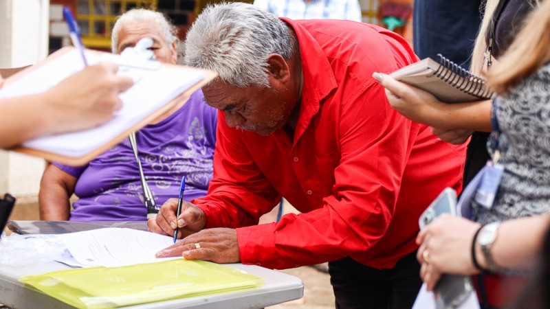 Um homem indígena curvado sobre uma mesa assina um documento com uma caneta.