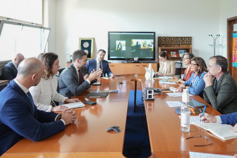Foto em sala de reunião, de paredes brancas. Leite aparece sentado junto à mesa retangular de madeira, à esquerda, e gesticula enquanto fala. À sua frente está Maria. Há outras pessoas sentadas na sala, e uma TV grande ao fundo.