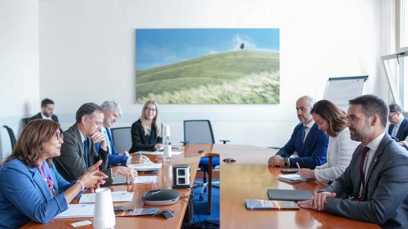 Foto em sala de reunião, de paredes brancas. Leite aparece sentado junto à mesa retangular de madeira, à direita. à sua frente está Maria, que fala. Há outras pessoas sentadas na sala.