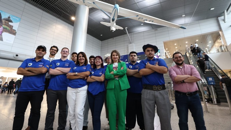 Foto posada no saguão do aeroporto. Em torno de dez estudantes, de camisetas azuis, posam sorrindo para a câmera algo de alguns adultos.