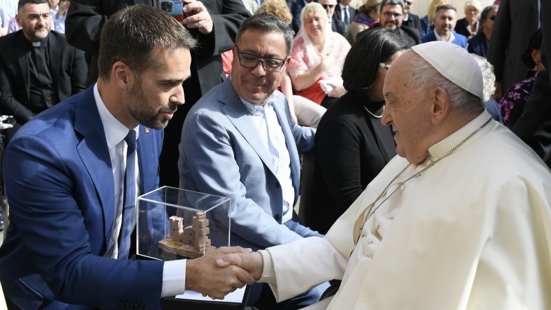 O governador Eduardo Leite, do lado esquerdo da foto, aperta a mão do Papa Francisco, posicionado do lado direito. Leite segura uma escultura das ruínas de São Miguel das Missões, com a qual presenteará o pontífice. Ao fundo da foto, aparecem outras pessoas. 