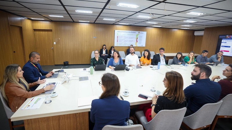 Foto de sala de reunião, com uma grande mesa retangular branca, junto da qual cerca de 15 pessoas estão sentadas.