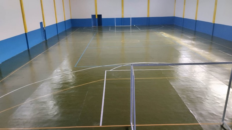 Foto geral de uma quadra poliesportiva. O espaço é fechado, o piso é verde-musgo, e há traves de gol de futebol instaladas.
