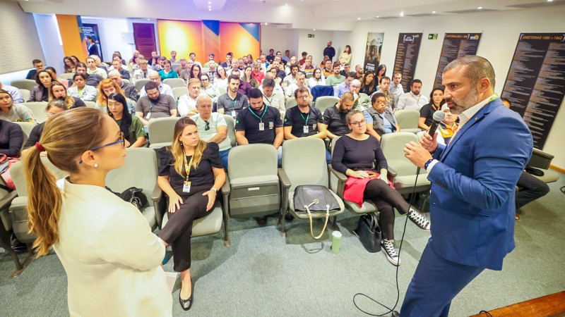 Foto tirada por detrás dos condutores do encontro, enquadrando a plateia sentada em cadeiras ao fundo. No primeiro plano, aparecem um mulher e um homem de pé, ambos de lado para a câmera. O homem fala ao microfone.