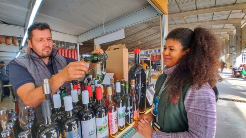 Foto num pavilhão da feira. Um produtor, em seu estande, serve cachaça para uma cliente, que sorri enquanto espera.