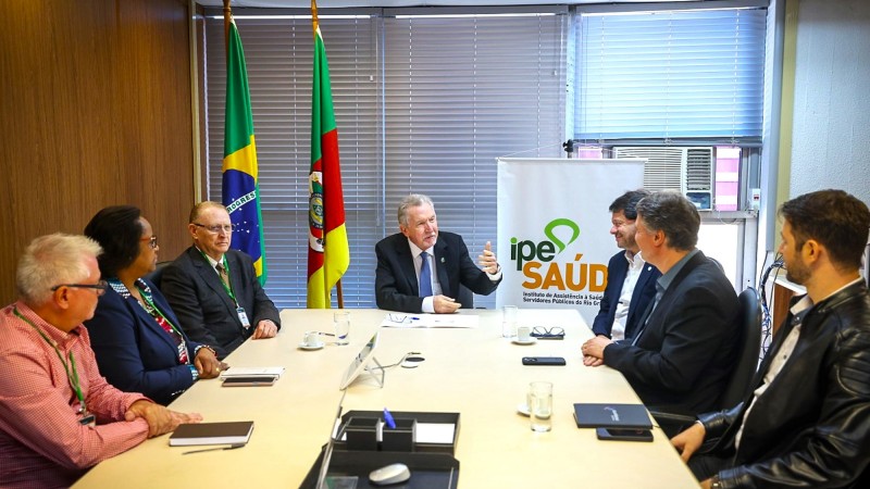 Imagem de pessoas sentadas conversando em uma mesa de reunião. Há um homem na cabeceira e dois homens e uma mulher do lado esquerdo e três homens do lado direito. No fundo, podem ser vistas uma bandeira do RS e uma do Brasil, além de um banner com a logomarca do IPE Saúde.