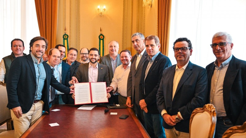 Gabriel segura o documento recém assinado para o setor Leiteiro rodeado por 12 homens. A foto é posada em um ambiente dentro do Palácio Piratini onde ocorreu a reunião.