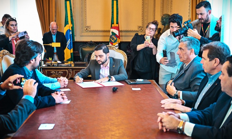 Imagem de Gabriel sentado à mesa de reunião assinando o decreto para o setor leiteiro. Há pessoas sentadas à mesa acompanhando o ato e outras, em pé, fazendo o registro audiovisual.