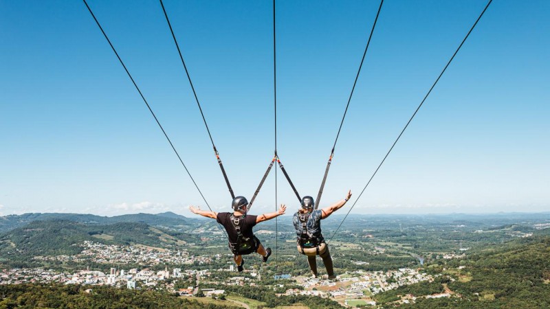 Imagem de duas pessoas de costas descendo de tirolesa em Feliz. Na imagem, a cidade pode ser vista do alto e à distância.