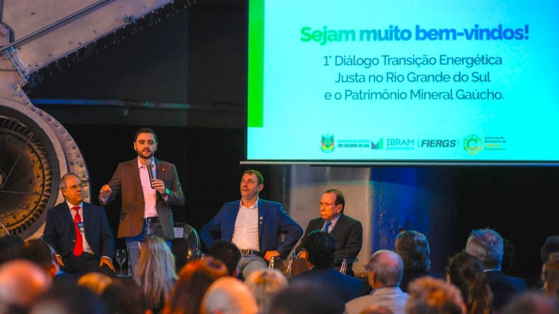 Gabriel falando ao microfone em pé, ao lado de outros três homens sentados que participam da abertura do evento. Há uma projeção mais à direita da imagem: Sejam muito bem-vindos. Primeiro diálogo Transição Energética Justa no Rio Grande do Sul e o Patrimônio Mineral Gaúcho. Uma parte da plateia pode ser vista de costas assistindo ao vice-governador.