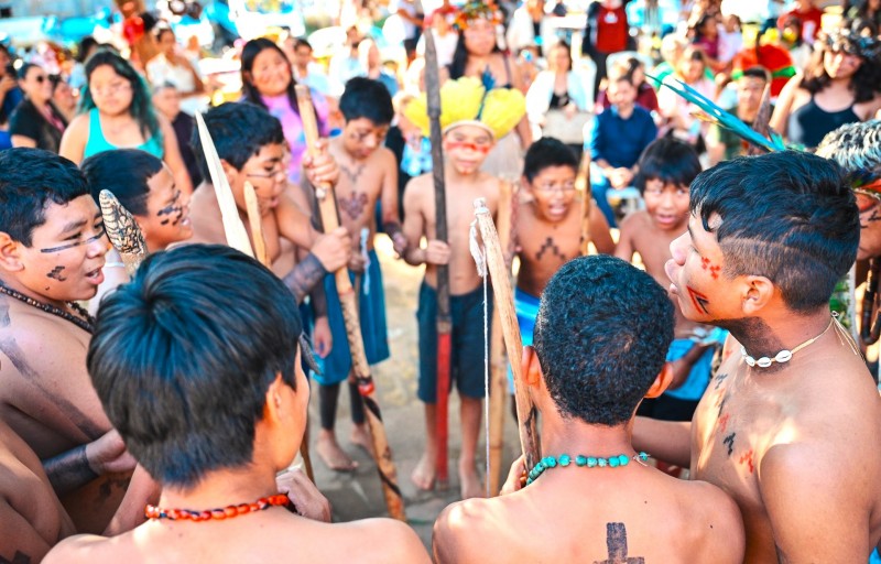 Círculo de crianças e adolescentes indígenas dançando com pintura corporal e segurando objetos indígenas durante a entrega da reforma na escola indígena Fag Nhin.