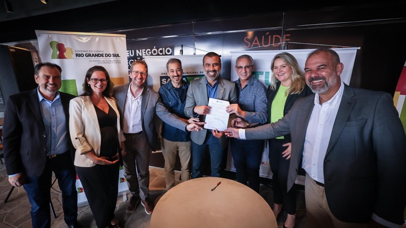 Foto posada, na qual estão seis homens e duas mulheres em pé atrás da mesa onde o documento foi assinado. Quatro homens seguram o documento recém-assinado.