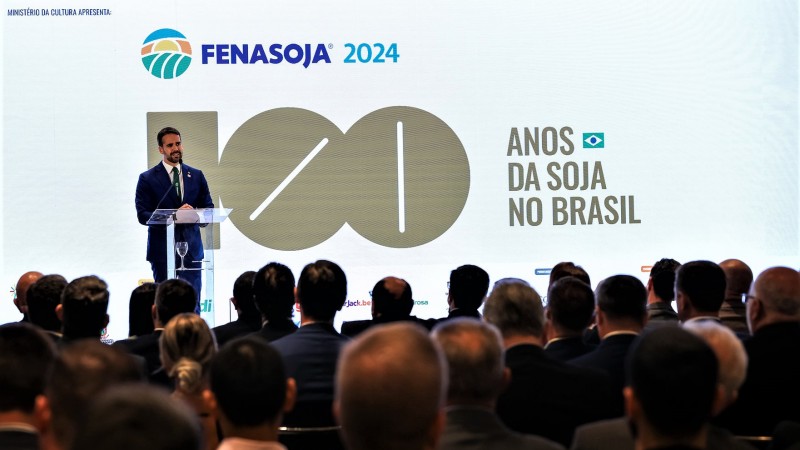 leite atrás de um púlpito falando ao microfone. A plateia é vista de costas e atrás de Leite há um imenso painel onde se lê. Fenasoja 2024, cem anos da soja no Brasil.