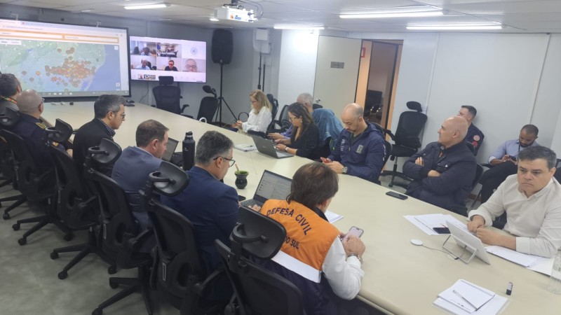 Foto geral de uma sala de reunião com uma grande mesa retangular branca. Em torno de 15 pessoas aparecem sentadas junto a ela. No fundo há um telão com o mapa do RS.