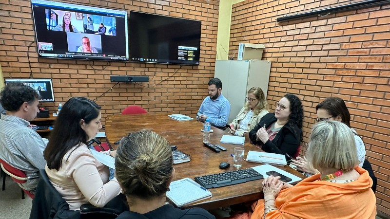 Reunião de alinhamento SES e GHC para tratar sobre atendimento a populações afetadas pelas enchentes. Oito pessoa em volta de uma mesa e diante de uma tela onde aparece mais três pessoas que participam à distância da reunião.