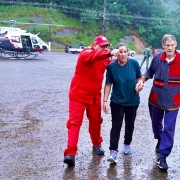 Casal acabando de ser resgatado em São Sebastião do Caí> eles caminha acompanhados por dois bombeiros. Ao fundo, pode ser visto o helicóptero.