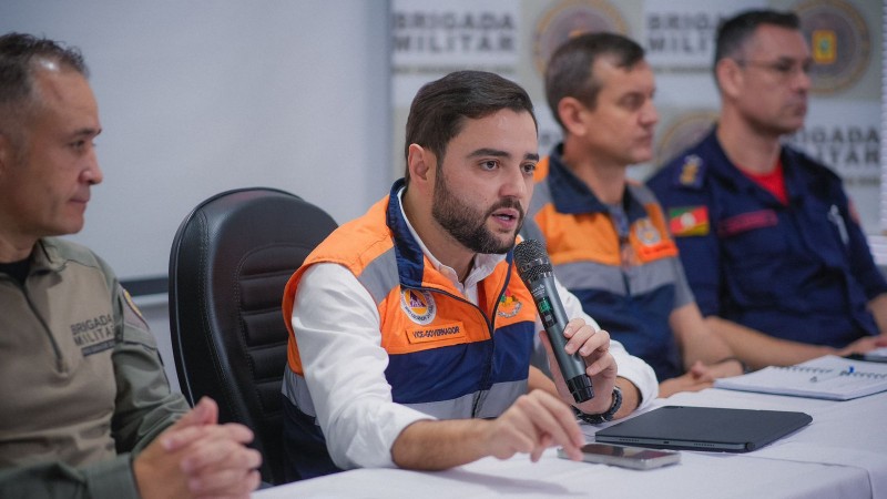 Foto de Gabriel concedendo entrevista coletiva. Ele aparece sentado, trajando colete laranja da Defesa Civil e falando ao microfone. Mais três homens aparecem sentados na imagem.