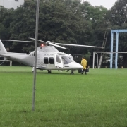Foto de um helicóptero branco pousado num gramado. Um homem de capa de chuva amarela se dirige a ele.
