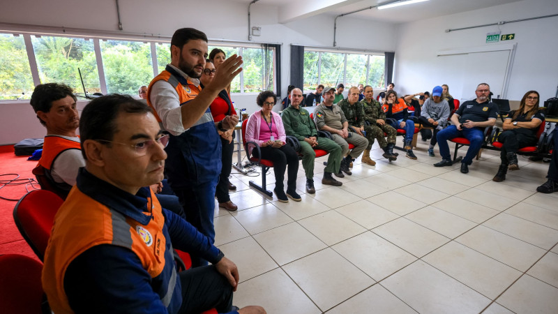 Foto de uma reunião numa sala de paredes e piso branco. Mais de 15 pessoas encontram-se sentadas em cadeiras dispostas em círculo. Gabriel está de pé, à esquerda do quadro, e gesticula enquanto fala.