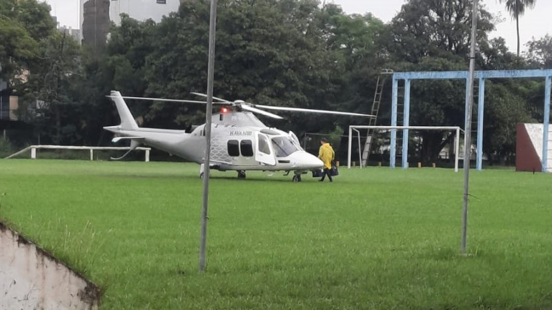 Foto de um helicóptero branco pousado num gramado. Um homem de capa de chuva amarela se dirige a ele.