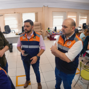 Gabriel conversando com outros três homens na instauração do gabinete de crise avançado da região Metropolitana. è uma sala grande onde outras pessoas estão trabalhando.